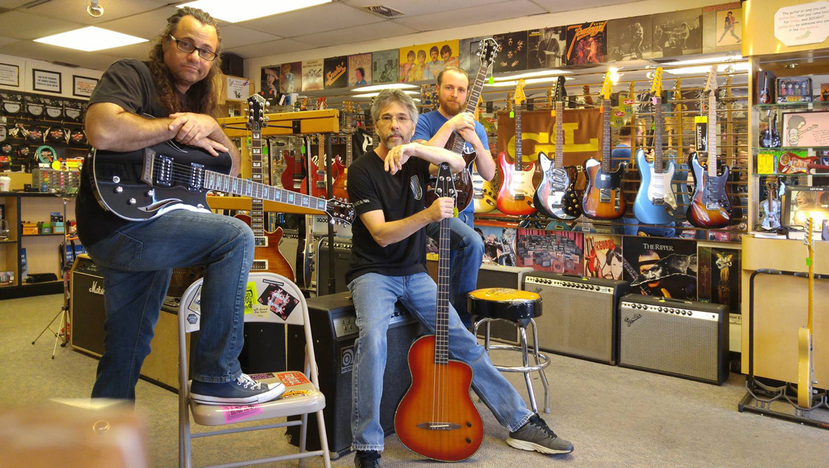 Stevie B's music store staff holding Michael Kelly Hybrid, Rick Turner Bass, and Patriot electric guitars
